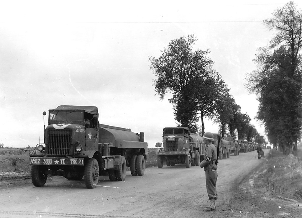 A column of US Army Quartermaster Truck Company tractor units and semi trailers pass a Military Policeman on a tree lined road showing clear bumper markings.