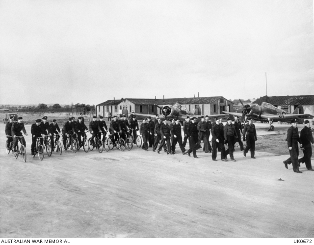 A group of uniformed people walking and riding bicycles past a group of buildings and single engined aeroplanes.